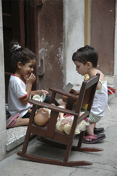 Deux jeunes enfants (une fille et un garçon) derrière une chaise à bascule jouent sur le seuil d'une maison. Le garçon manipule attentivement un objet tandis que la fillette rit avec sa main devant la bouche. Il y a deux poupées sur la chaise.