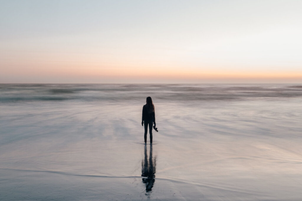 Personnage se trouvant debout sur l'estran de la plage. Crépuscule, on devine le coucher de soleil sur la droite.